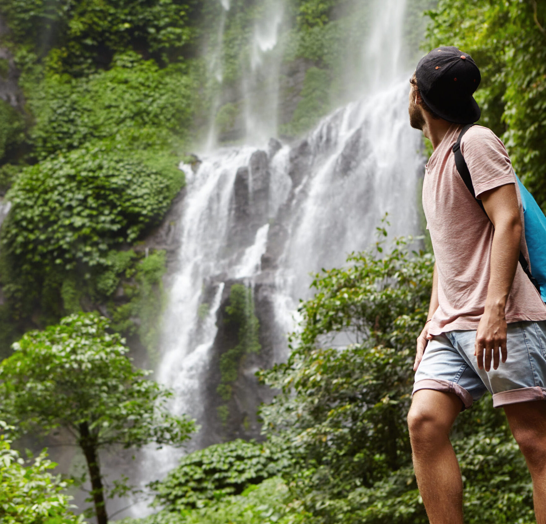 People, wildlife and adventure concept. Fashionable young adventurer with backpack contemplating waterfall landscape. Male tourist enjoying beautiful nature around him during his journey in rainforest